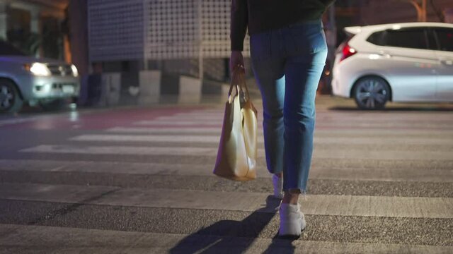 Rear view of Confidence young beautiful Asian woman holding leather bag crossing city street crosswalk at night. Pretty girl enjoy walking in the city with looking illuminated night lights