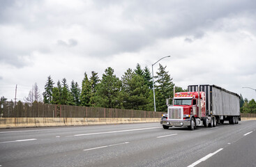 Red classic big rig semi truck tractor with vertical pipes and chrome accessories transporting cargo in covered bulk semi trailer moving on the wide multiline highway road