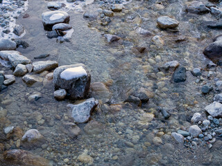 Clear mountain water flows out from under the ice. Small stones are dusted with snow