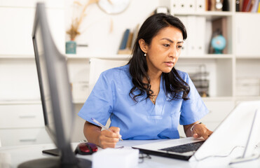 Latin American doctor writing notes and reading something on laptop at her office