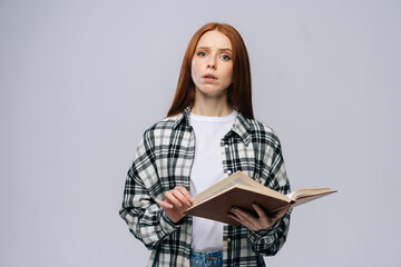 Portrait of young woman college student holding opened books and looking at camera on isolated gray background. Pretty redhead lady model wearing casual clothes emotionally showing facial expressions