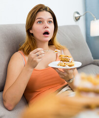 Cheerful young female eating sweet cake at home on cozy sofa