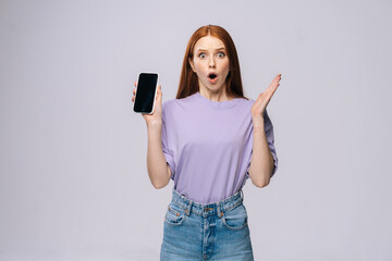Fototapeta premium Shocked young woman wearing stylish casual clothes holding cell phone with black empty mobile screen. Pretty lady model with red hair emotionally showing facial expressions in studio, copyspace.