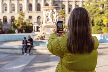 Photo on the phone. A young tourist in  yellow jacket takes pictures of sights in the city. View from the back.