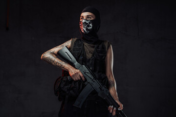 Fototapeta na wymiar Tattooed and seductive woman in black armour and weared with mask poses in dark background holding assault rifle.
