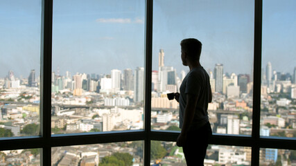 Dreaming european man stands by the window with great views and drinks hot coffee or tea, enjoying beautiful morning feeling happy