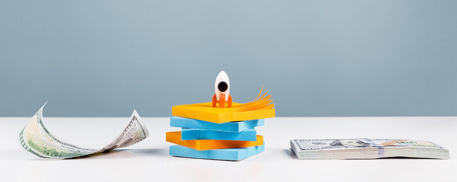 Business Or Education Growth Development And Success Concept Banner. Rocket And Money Stack On White Office Or Home Desk With Blue Background. Innovative Thinking. Copy Space