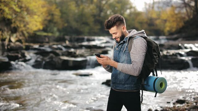A man is standing on the shore of a mountain river and texting on a smartphone. Hiking in the mountains. 4K.