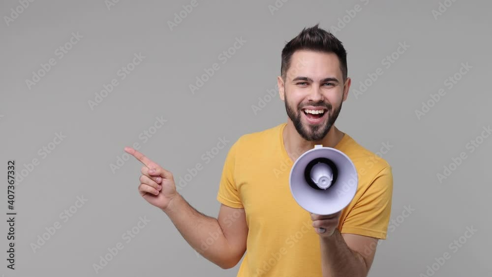 Wall mural Cheerful excited crazy bearded young man 20s years old in yellow casual t-shirt isolated on grey color background studio. People lifestyle concept. Screaming in megaphone pointing index finger aside