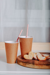 Heart-shaped cookies on round wooden cutting board. Saint Valentine's cookies in shape of heart and craft coffee cups on white background. Two paper coffee to go with tubes.