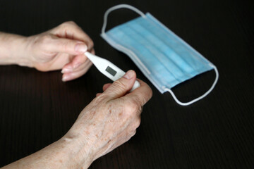 Elderly woman measuring body temperature, concept of fever, coronavirus symptoms. Digital thermometer in wrinkled female hands on medical mask background