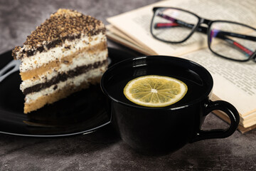 Tea with lemon and cake in black dishes glasses on a book on a gray background