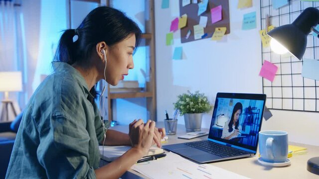 Asia businesswoman using laptop talk to colleagues about plan in video call meeting at living room. Working from house overload at night, remotely work, social distancing, quarantine for coronavirus.