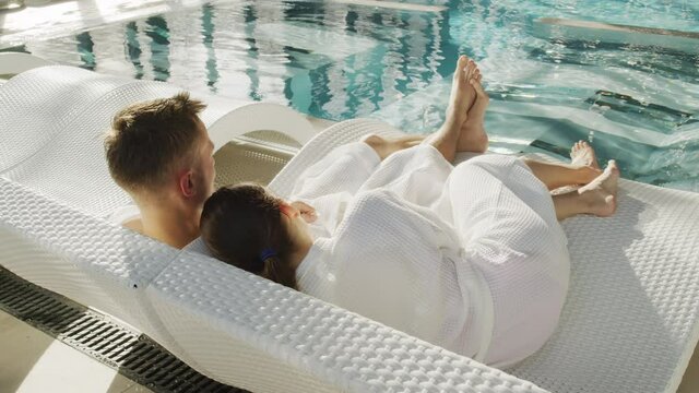 Back-view high-angle slowmo of loving couple in white bathrobes chilling on loungers at poolside looking at blue transparent water enjoying moments together
