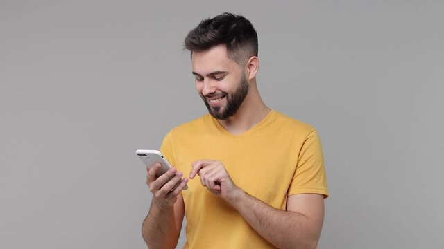 Smiling cheerful bearded young man 20s years old wearing yellow casual basic t-shirt isolated on grey color wall background studio. People lifestyle concept. Using mobile cell phone typing sms message