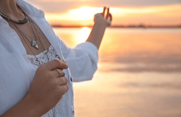 Young woman holding amulet near river at sunset, closeup view with space for text. Nature healing...