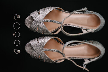 Silver shoes, earrings and rings, composition isolated on the black background. Accessory bridal. Footwear. Wedding rings and sandal. Shoe with crystal. Engagement. Close up. Flat lay. Top view.