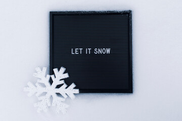 Black letter board with white words "Let it snow" with big shiny decorative snowflake, on the background of snowy road.
