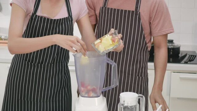 Happy Asian beautiful young family couple husband and wife making fresh smoothie in kitchen together at home. The man and woman pour the chopped apples into a juice blender. Slow motion