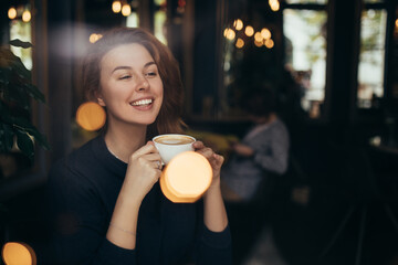 Young woman sitting in a cozy coffehouse drinking coffee near window.