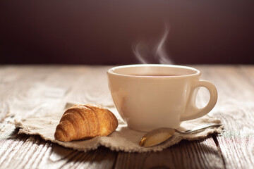 Steaming tea on a wooden table with a croissant lying next to it.