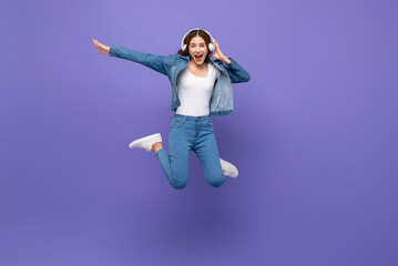 Fun energetic young Asian woman jumping and listening to music on headphones isolated on purple studio background