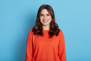 Obraz premium Positive joyful young beautiful female with dark hair wearing casual clothing, looking at camera and pleasantly smiling, wearing orange sweater, standing isolated over blue background.
