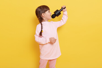 Side view of singing little girl wearing pale pink attire and having pigtails hairstyle, child holding microphone and performing against yellow background.