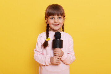 Cute little girl with microphone posing isolated on yellow color background, singing or telling...