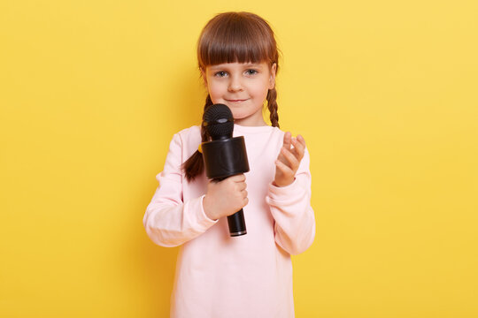 Beautiful little girl with microphone performing, looking at camera with charming smile, raising hand, looks a bit shy, child model posing isolated over yellow background.