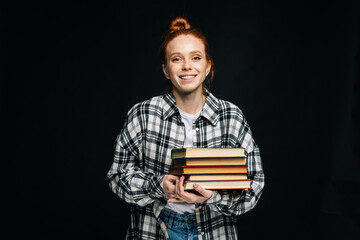 Cheerful young woman college student holding book and looking at camera on isolated black background. Pretty redhead lady model wearing casual fashion clothes emotionally showing facial expressions.