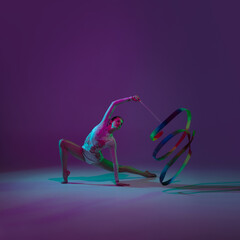 Rainbow. Young female athlete, rhythmic gymnastics artist dancing, training with tape isolated on purple studio background in neon light. Beautiful girl practicing with equipment. Grace in performance