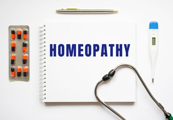 HOMEOPATHY is written in a notebook on a white table next to pills and a stethoscope.
