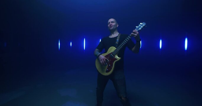 Male rocker playing guitar and singing song in dark room