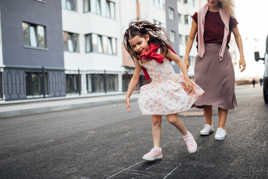 Happy Little Girl Playing Hopscotch With Her Mother On Playground Outdoors. A Child Plays With Her Mom Outside. A Kid And A Young Woman Play Hopscotch Drawn On The Pavement Outside.