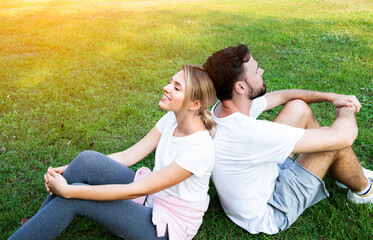 Romantic couple of young people keep eyes closed and look relaxed while sitting with back to each other on grass in park. Couple man and woman sit back on green grass outdoors.