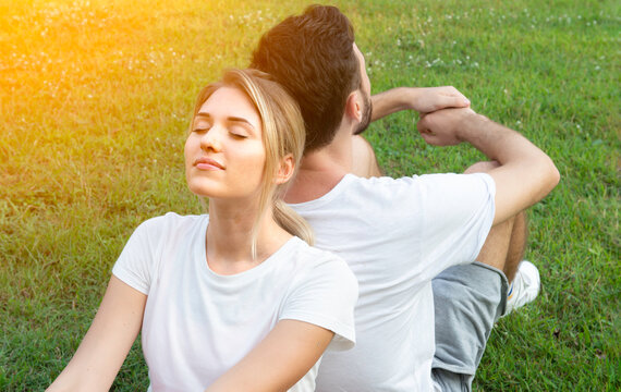 Romantic couple of young people keep eyes closed and look relaxed while sitting with back to each other on grass in park. Couple man and woman sit back on green grass outdoors. - Powered by Adobe