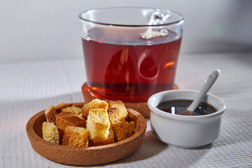 Square toasted pieces of homemade delicious rusk, hardtack, Dryasdust, zwieback, Liquid honey in a saucer and black tee in a cap on a white tablecloth.