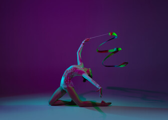 Fototapeta na wymiar Wave. Young female athlete, rhythmic gymnastics artist dancing, training with tape isolated on purple studio background in neon light. Beautiful girl practicing with equipment. Grace in performance.