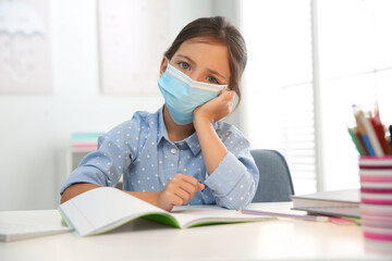 Distance learning, online studying at home due to Covid-19 pandemic. Sad girl in protective mask doing homework in room