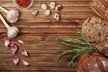Garlic, cereal bread, rosemary, quail eggs and a plate of paprika on a wooden table
