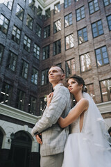 Fototapeta premium A cute curly-haired bride in a white dress with a long veil hugs the groom in a gray suit against the background of a multi-storey brick building. Wedding portrait of newlyweds in love.