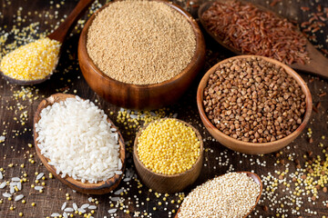 White and red rice, buckwheat, amaranth seeds, corn groats, quinoa and millet in bowls on a brown wooden table. Gluten-free cereals. Closeup