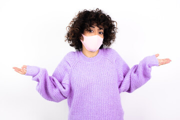 Clueless young beautiful caucasian woman wearing medical mask standing against white wall shrugs...