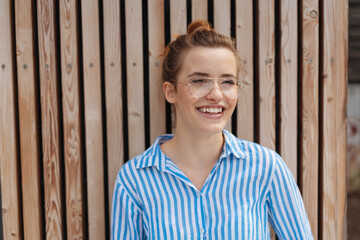 young laughing woman with glasses leans against a wooden wall and looks to the side