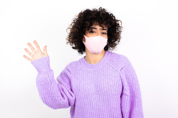 young beautiful caucasian woman wearing medical mask standing against white wall Waiving saying hello happy and smiling, friendly welcome gesture.