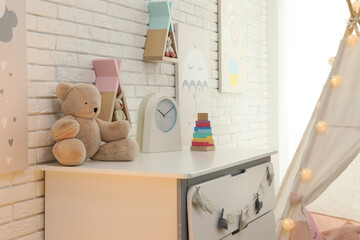 Toys and clock on chest of drawers near white brick wall in playroom. Interior design