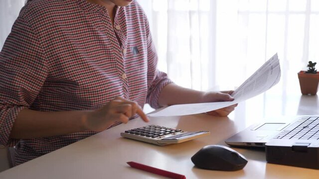 Anonymous young asian woman sitiing at a desk near white curtain, grabbing paper, using calculator to sum balance sheet, checking or validating. Indoors, side view, working from home concept