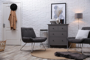Stylish room interior with grey chest of drawers and chairs near white brick wall
