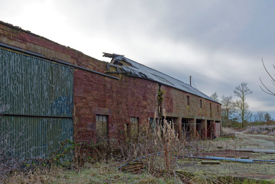 A ruined stone Farm Barn with the slate Roof fallen in, and bits of equipment lying on the hardstanding next to the machinery sheds.
